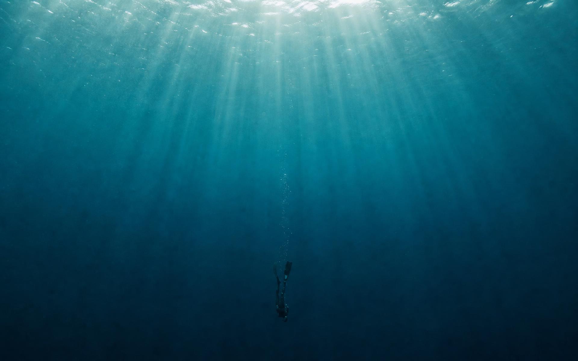 A lone freediver descending through cathedral god-rays in vast deep-teal water